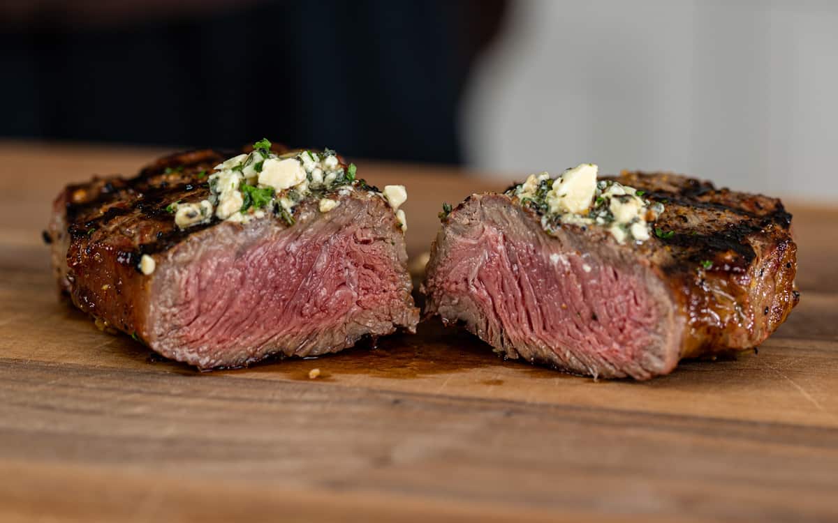 medium rare grilled rib eye steak topped with herb butter, cut in half on wood cutting board