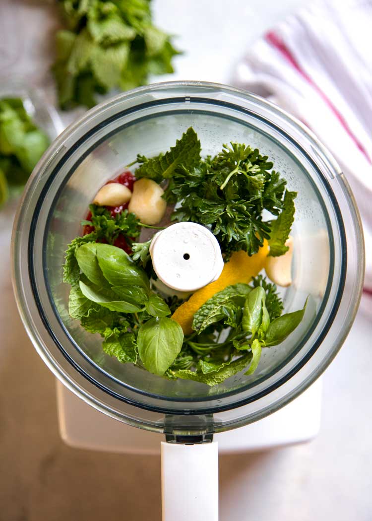 overhead photo of fresh mint, parsley, garlic, and lemon zest in food processor bowl