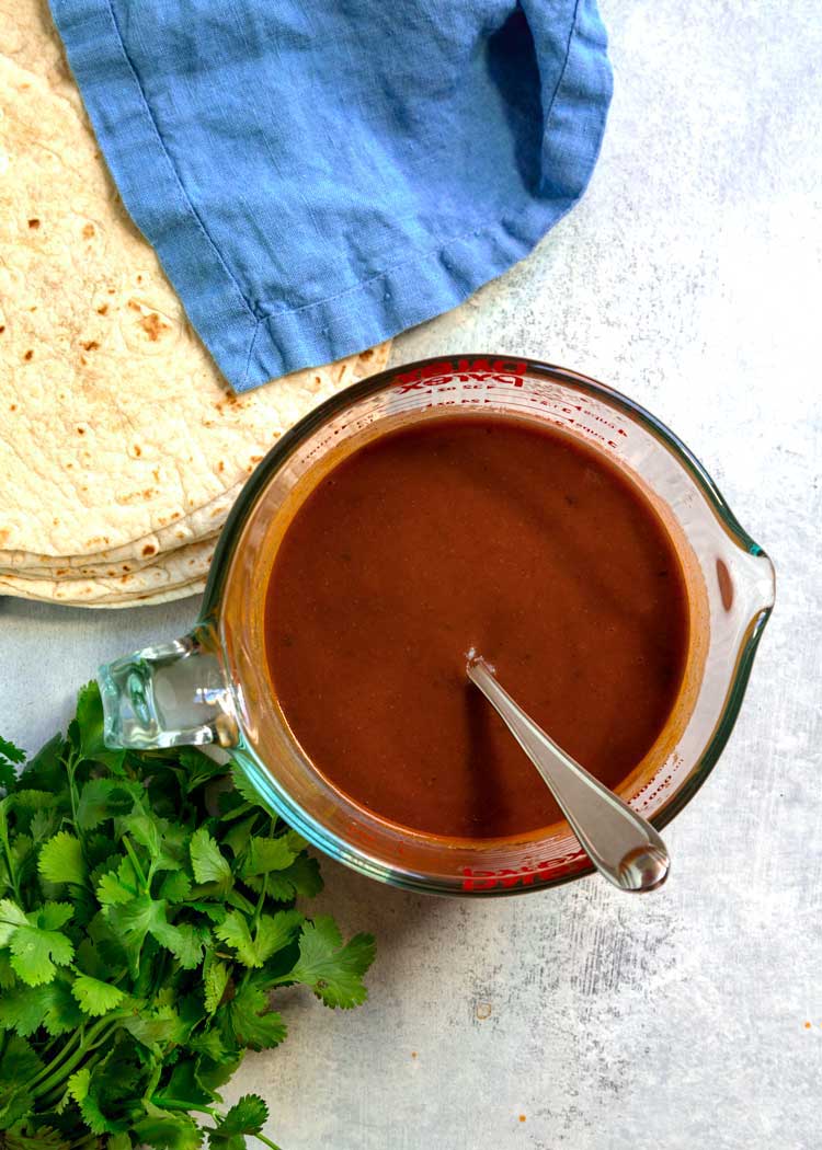 a measuring cup of Mexican red sauce next to tortillas and cilantro