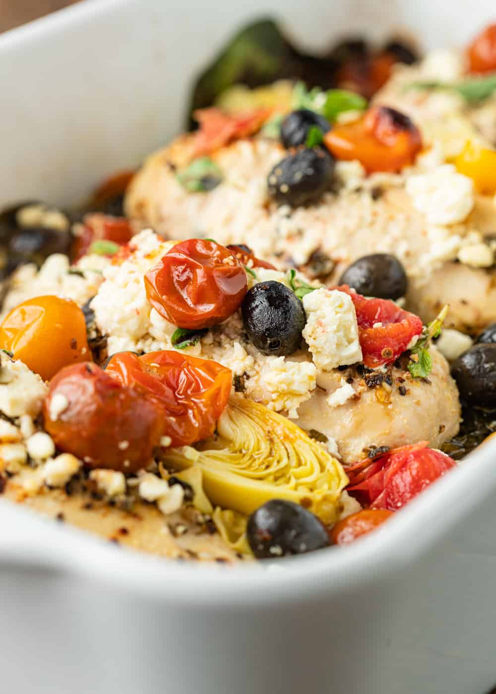 close up photo of baked chicken breasts topped with Greek olives, tomatoes, and artichoke hearts