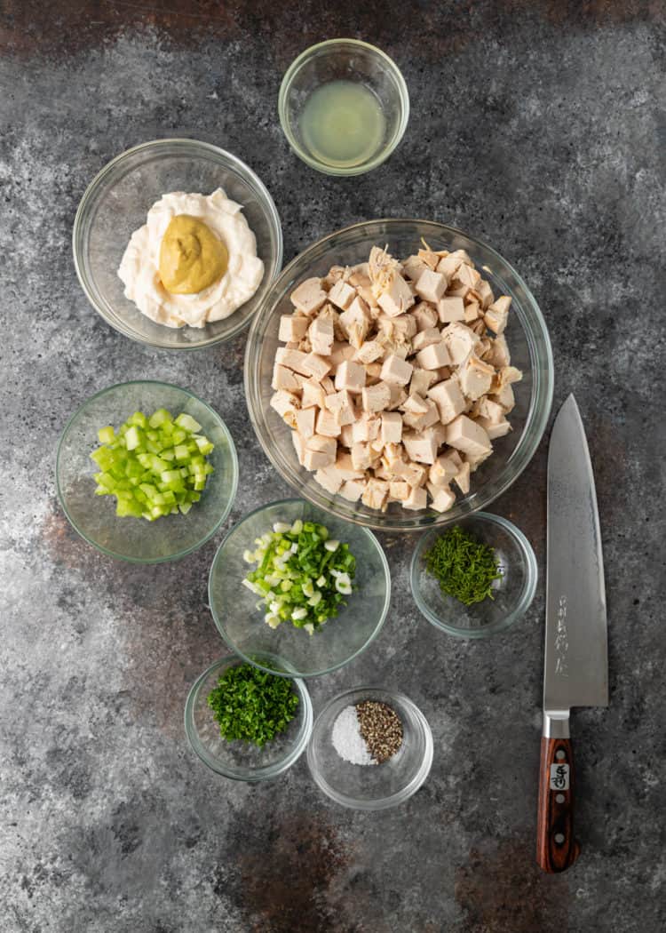 chicken salad ingredients in glass bowls on counter: chicken, celery, scallions, mayo, mustard and spices