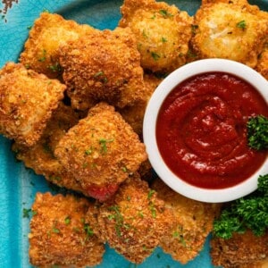 overhead photo of a white bowl of marinara surrounded by fried breaded ravioli and fresh parsley on a blue platter