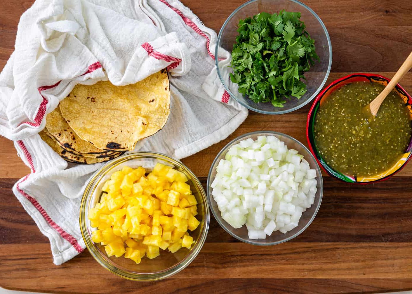 bowls of diced pineapple, diced onion, fresh cilantro, and green chili sauce next to corn tortillas wrapped in a white kitchen towel