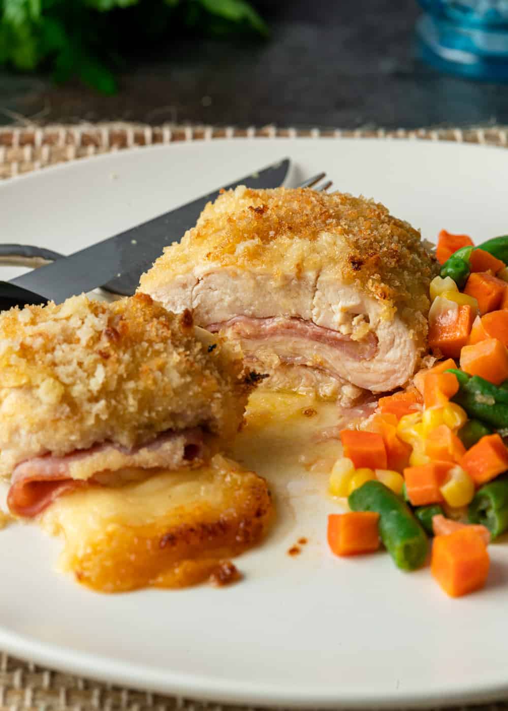 baked chicken cordon bleu on white dinner plate with mixed vegetables, fork and knife