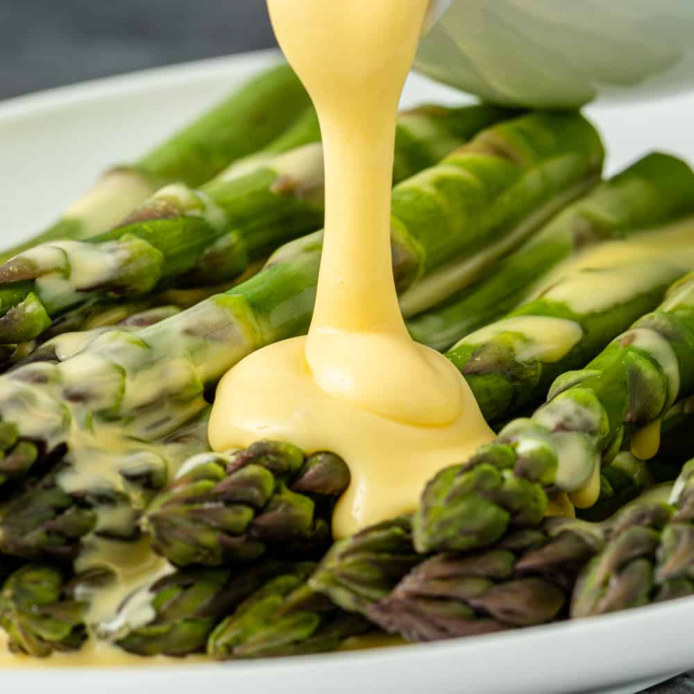 creamy hollandaise sauce being poured over steamed asparagus spears