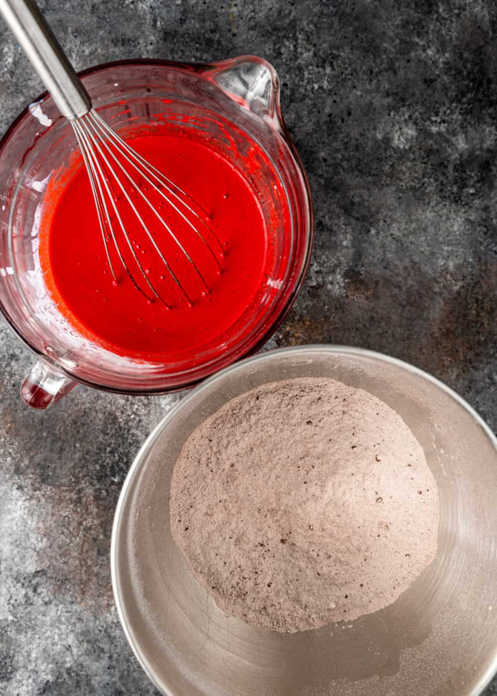 overhead shot of mixing bowl with flour next to measuring cup filled with red liquid and a whisk