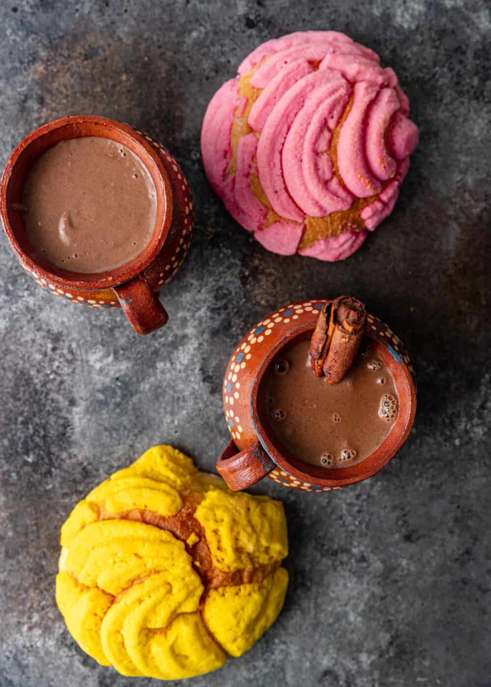 pink and yellow Mexican pastries next to 2 mugs of creamy champurrado