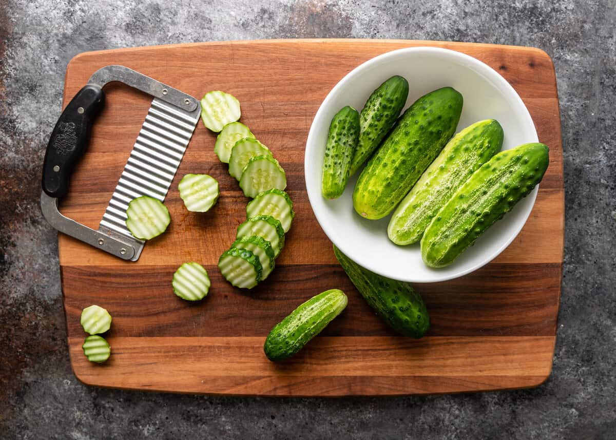 overhead: a bowl of cucumbers on a wooden cutting board with sliced cucumbers and a crinkle cutter