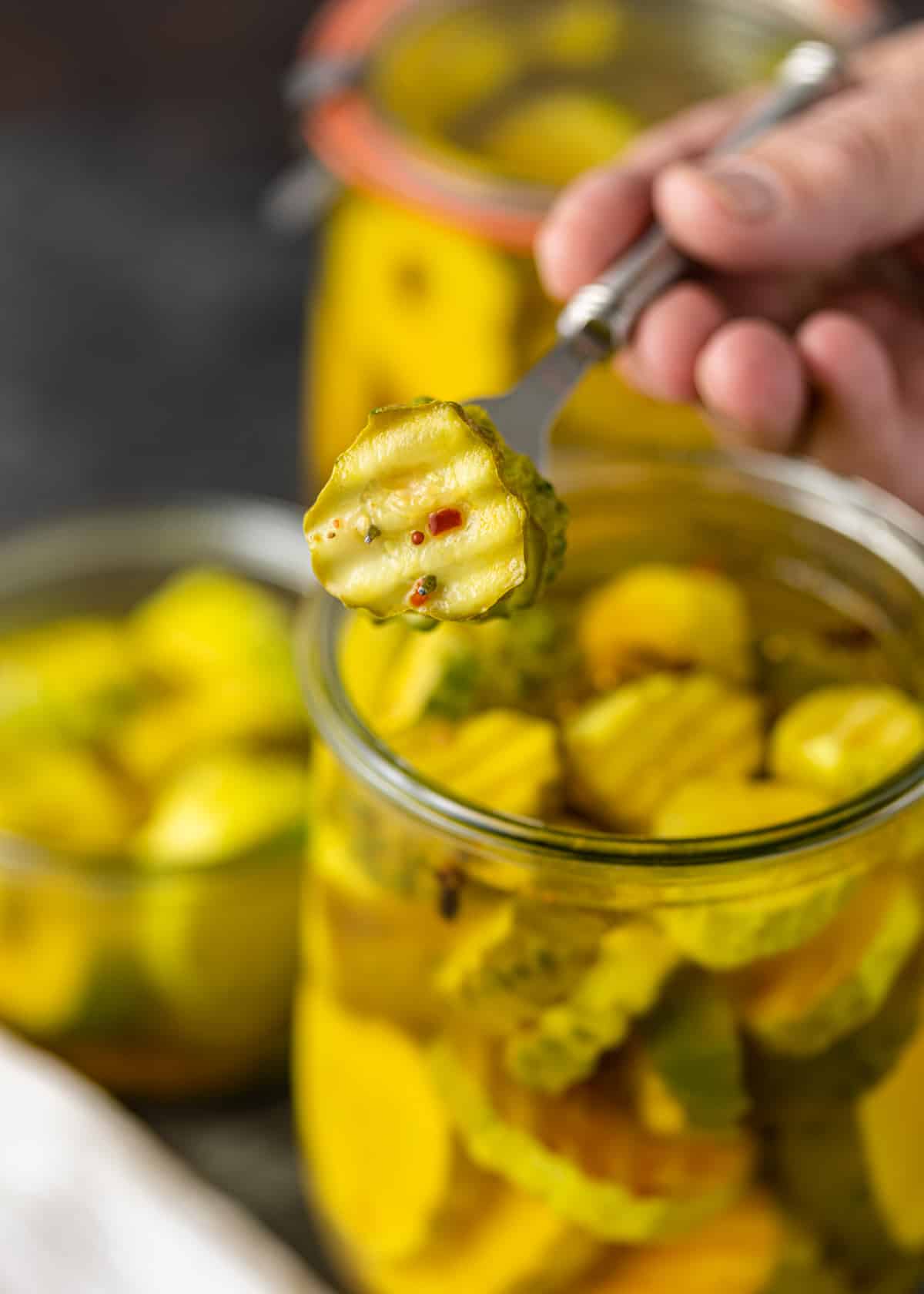 closeup: a fork lifting a bread and butter pickle out from a jar with more pickles