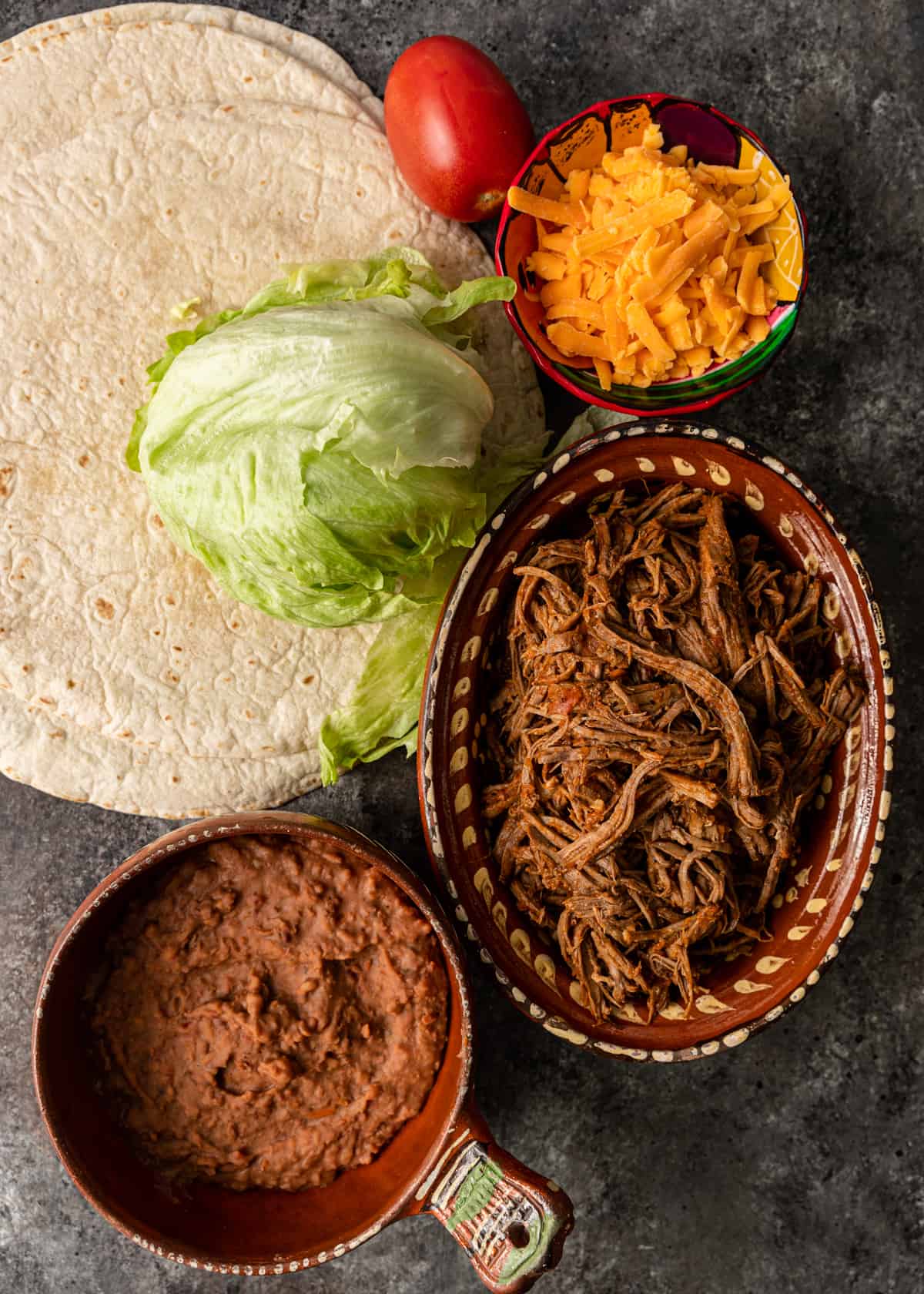 Top view of ingredients needed for shredded beef chimichangas including tortilla, shredded cheese, tomato, beef, refried beans, and lettuce.
