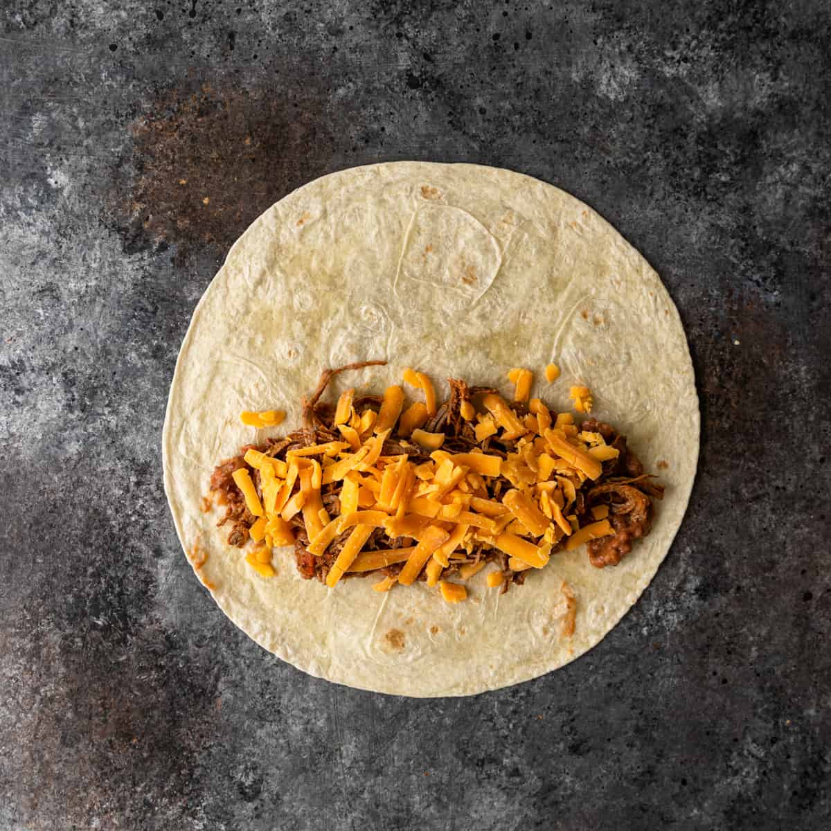 Top view of a tortilla with refried beans, shredded beef, and cheddar cheese.